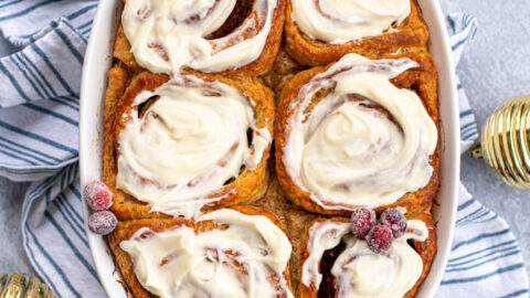 Overhead view of a white baking dish full of gingerbread cinnamon rolls topped with cream cheese frosting and garnished with sugared cranberries next to gold Christmas ornaments.