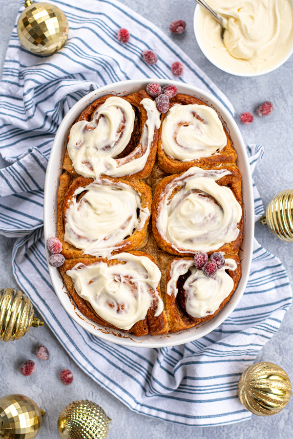 Overhead view of a white baking dish full of gingerbread cinnamon rolls topped with cream cheese frosting and garnished with sugared cranberries next to gold Christmas ornaments.