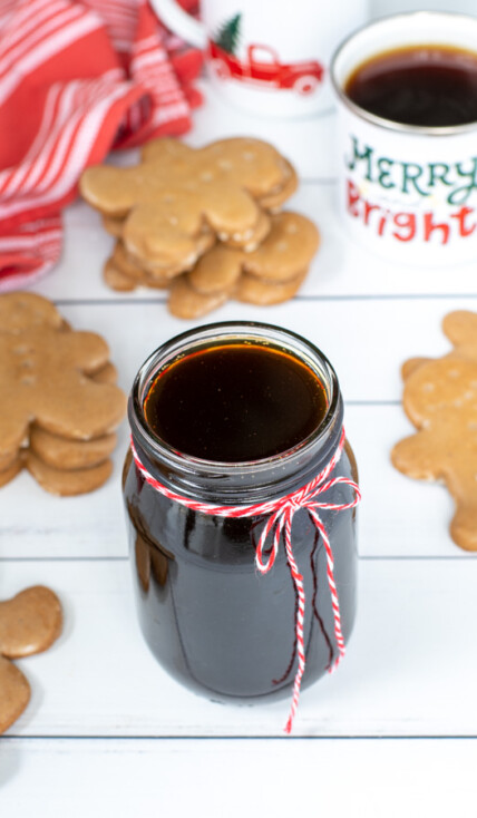 A glass jar of gingerbread simple syrup with a red and white bow on a table next to gingerbread cookies and mugs of coffee.