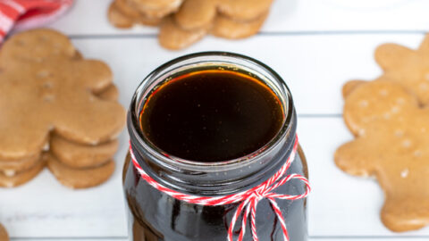 A glass jar of gingerbread simple syrup with a red and white bow on a table next to gingerbread cookies and mugs of coffee.