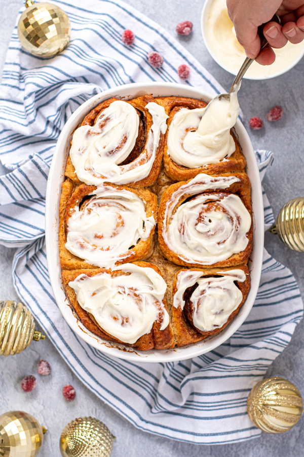 A hand with a spoon putting cream cheese frosting onto a freshly baked cinnamon roll.
