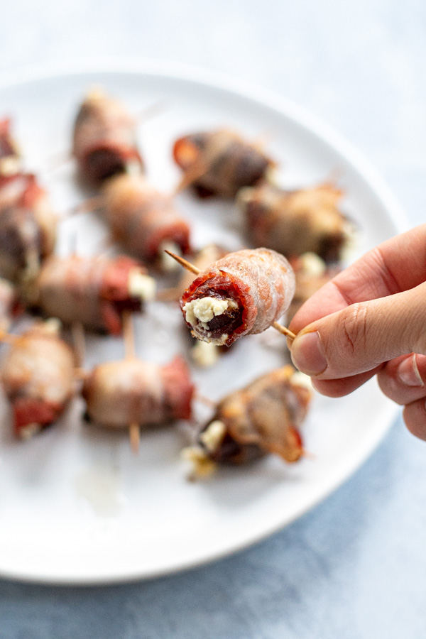 A hand holding a toothpick with a bacon wrapped date showing melted goat cheese coming out of the middle.
