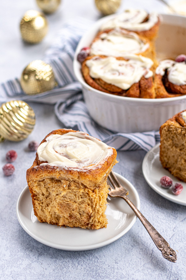 A fresh gingerbread cinnamon roll on a small white plate topped with cream cheese frosting.