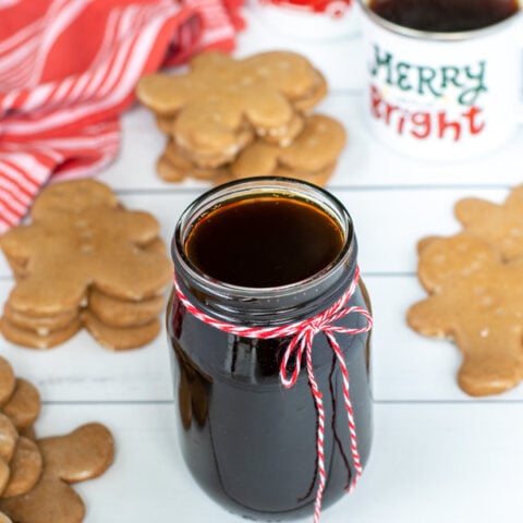 A glass jar of dark colored simple syrup sit-in non a table next to mugs of coffee and stacks of gingerbread cookies.