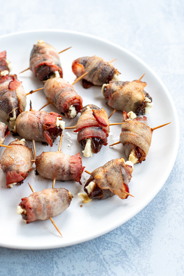 Side angle of bacon wrapped dates on a white plate showing goat cheese melting out and held together by toothpicks.