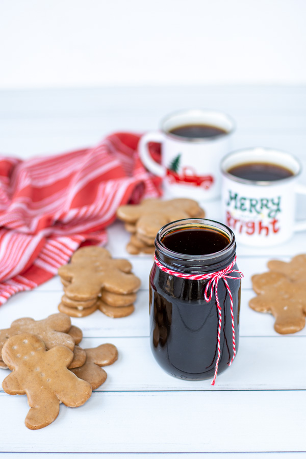 A glass jar of gingerbread simple syrup tied with a red and white string on a tale next to stacks of gingerbread cookies and mugs of coffee.