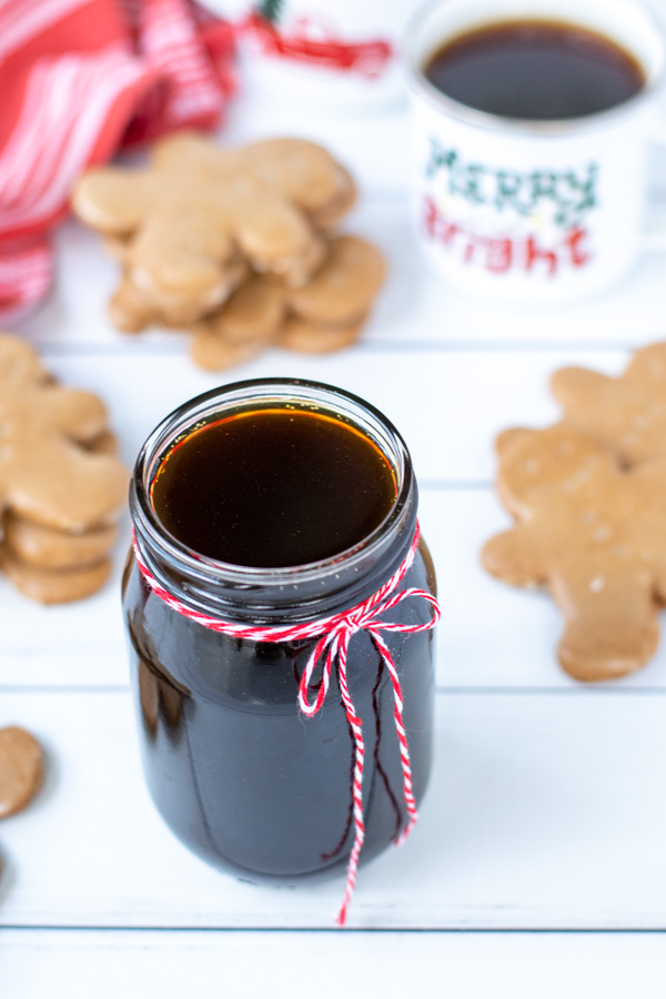 A styled shot of gingerbread syrup in a glass jar tied with a red and white string sitting next to stacks of gingerbread cookies and a mug of coffee.