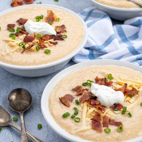 Three white bowls at an angle on a table filled with baked potato soup topped with cheese, bacon, greens onions, and a sour cream dollop.