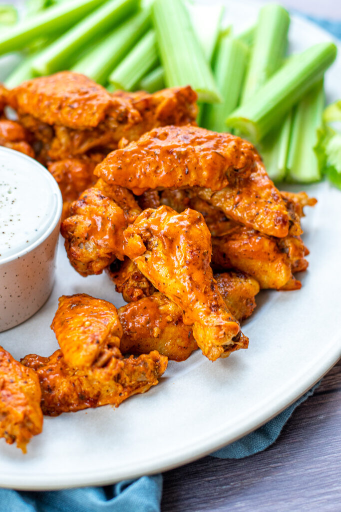 Stack of saucy hot wings on a white platter next to celery with a chicken crummier sitting front and center.