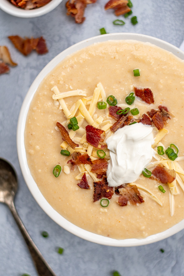 Close up of a bowl of baked potato soup with the toppings of shredded gouda cheese, green onions, crumbled bacon, and sour cream.
