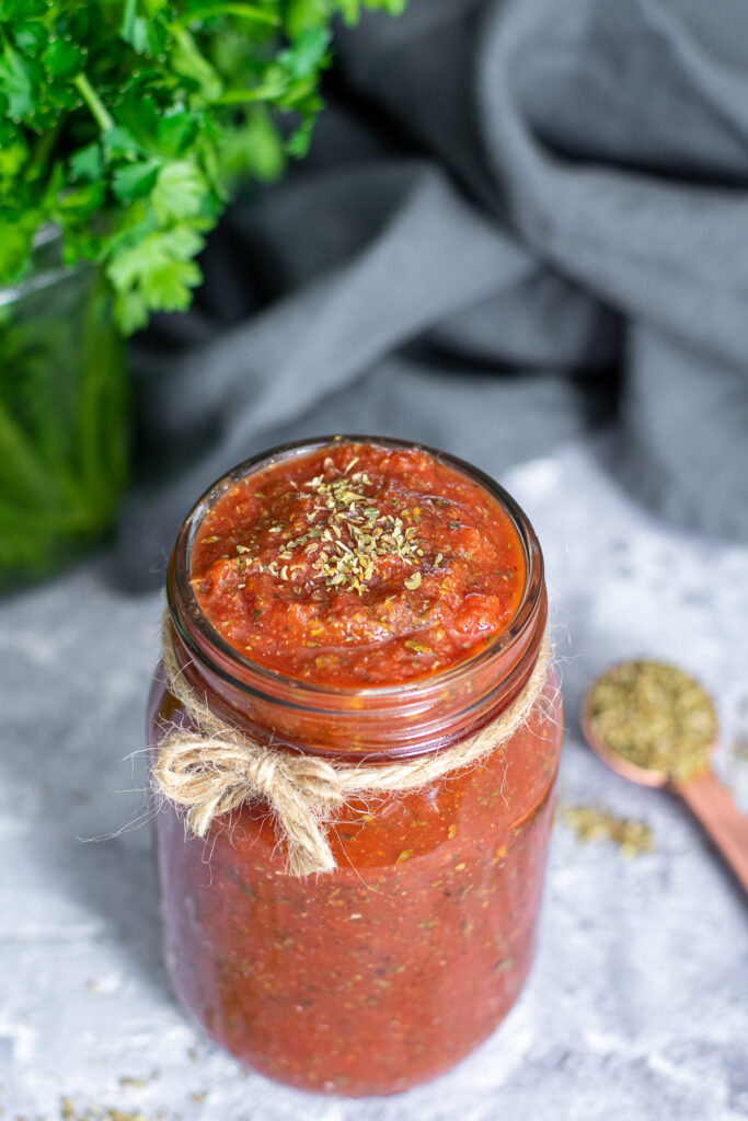 Close up of the open top of a glass jar filled to the brim with pizza sauce, topped with a little sprinkle of dried oregano.
