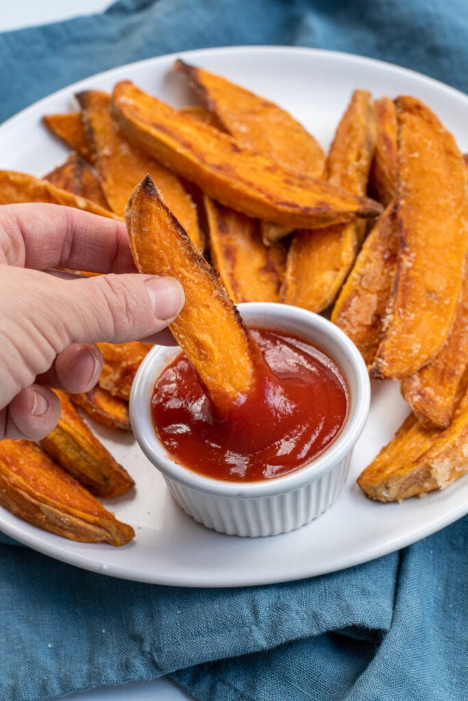 Hand dipping a crispy sweet potato wedge into ketchup.
