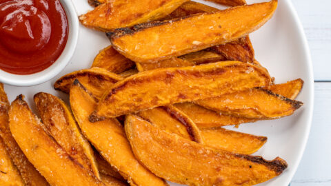 Close up of a white plate sitting on blue towel the plate in piled high with crispy sweet potato wedges and a little cup of ketchup.