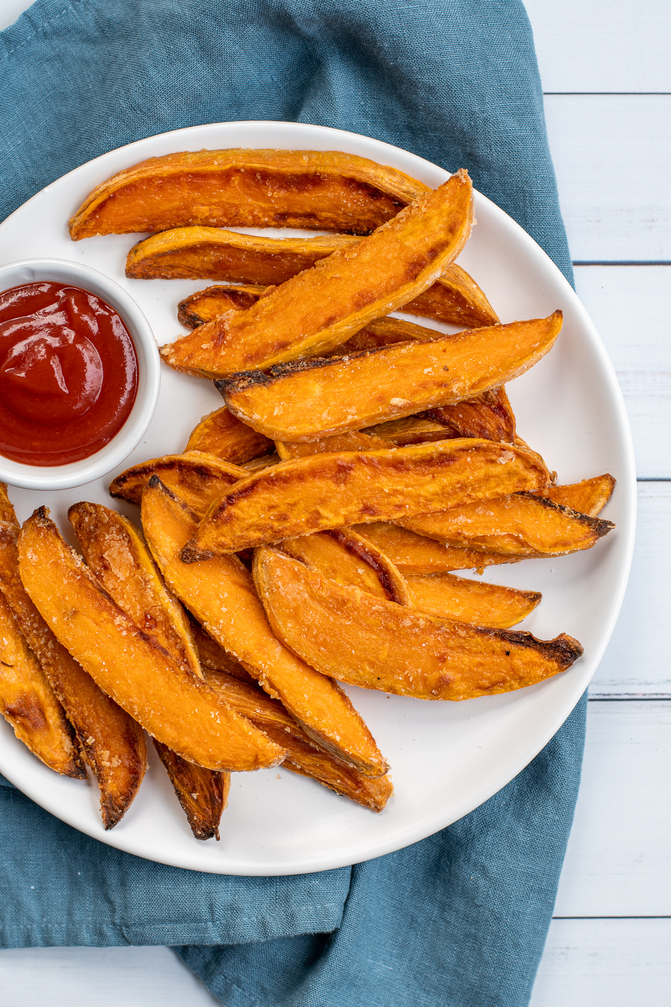 Close up of a white plate sitting on blue towel the plate in piled high with crispy sweet potato wedges and a little cup of ketchup.