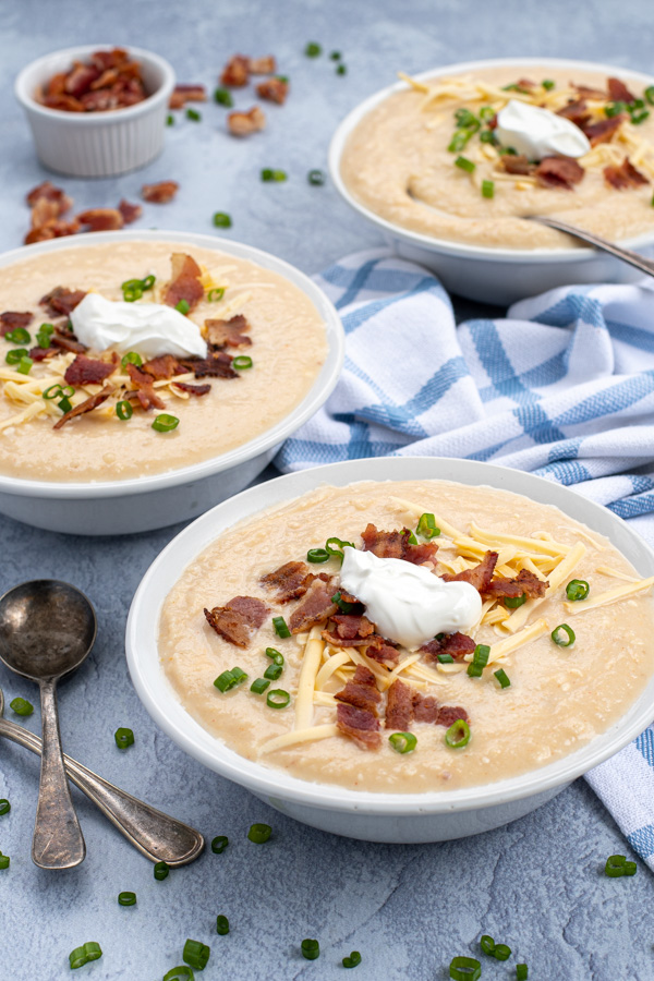 Instant Pot Loaded Baked Potato Soup in three bowls shown at an angle topped with cheese, bacon, green onions, and sour cream they are sitting next to a pile of spoons.