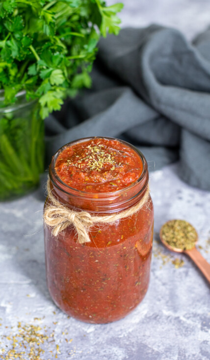 A glass jar with a little brown bow filled to the very top with 5 minute pizza sauce, topped off with a little pinch of dried oregano.