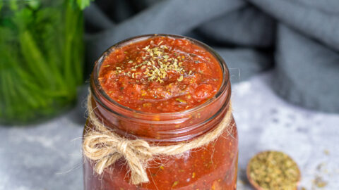 A glass jar with a little brown bow filled to the very top with 5 minute pizza sauce, topped off with a little pinch of dried oregano.
