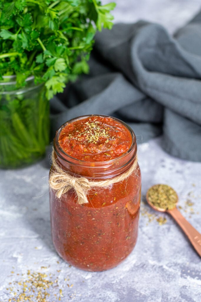A glass jar with a little brown bow filled to the very top with 5 minute pizza sauce, topped off with a little pinch of dried oregano.