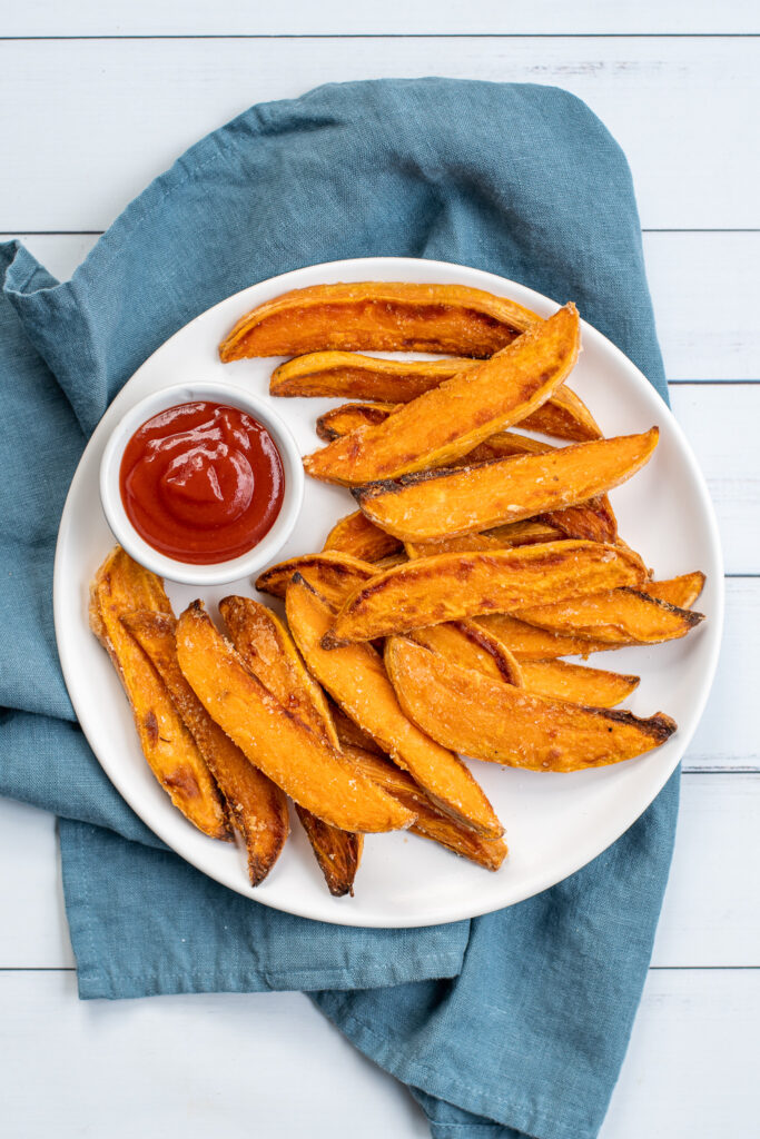 Overhead shot of a white plate sitting on blue towel the plate in piled high with crispy sweet potato wedges and a little cup of ketchup.