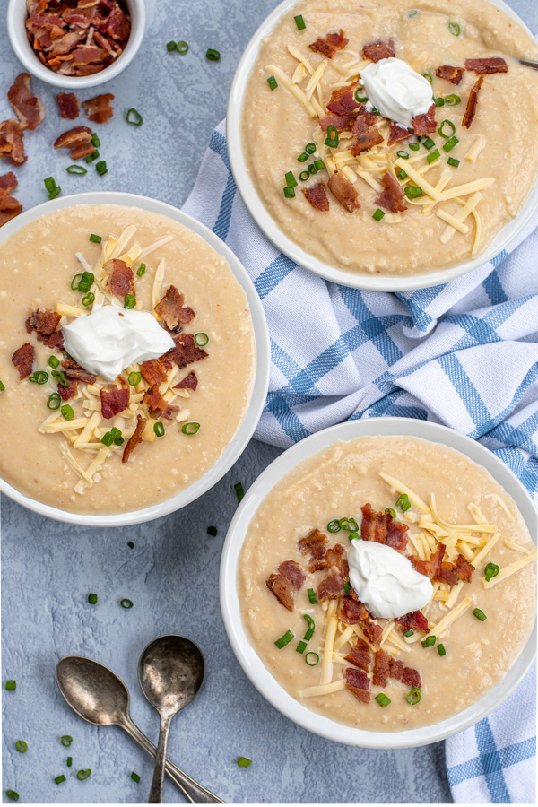 Flatly of three bowls filled with baked potato soup sitting on a gray background next to a towel, spilled bacon crumbles, and spoons. The soups are garnished with cheese, bacon, and green onions.