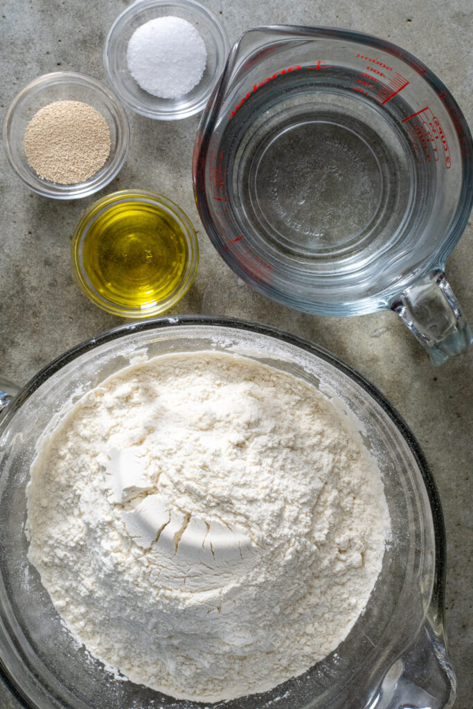 Ingredients including flour, water, olive oil, yeast, and salt laid out ready to be used to make pizza dough.