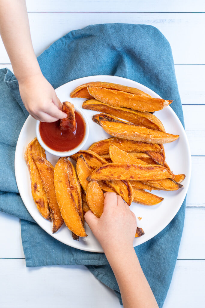 A white plate with pile of crispy sweet potato wedges with kid's grabbing the sweet potatoes and a little cup of ketchup.