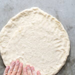 Pinterest Pin with text overlay 'Homemade Pizza Dough'. Image of pizza dough being. pressed into a crust shape on a gray counter.