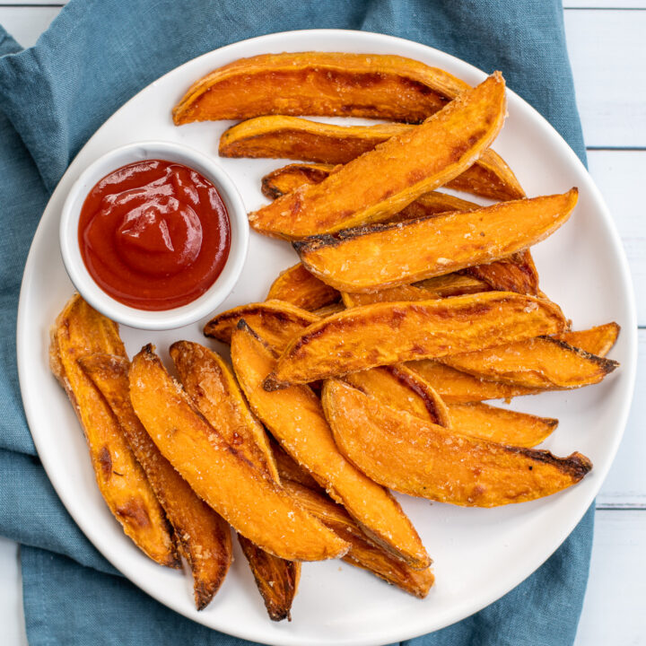 A white plate sitting on blue towel the plate in piled high with crispy sweet potato wedges and a little cup of ketchup.