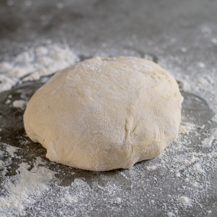 A ball of pizza dough covered on flour sitting on a flour covered countertop.