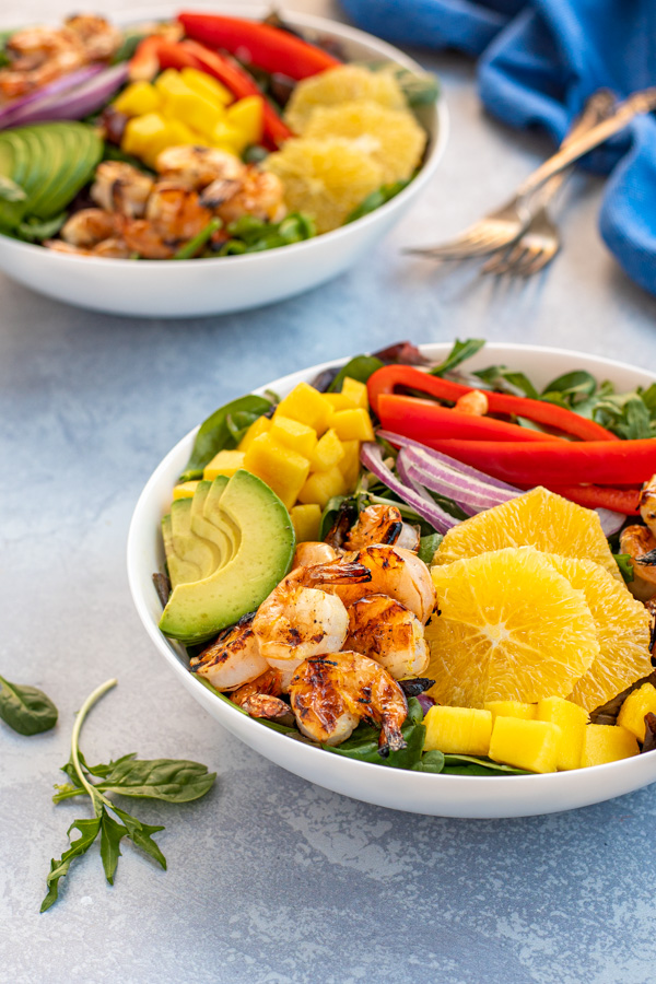 Two large bowls of grilled shrimp on green lettuce with avocados, oranges, mango, and bell pepper.