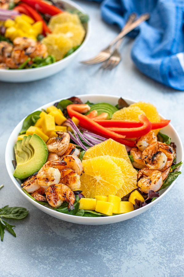A large white bowl on a table with mixed greens topped with grilled shrimp, avocado slices, oranges, and diced mango.