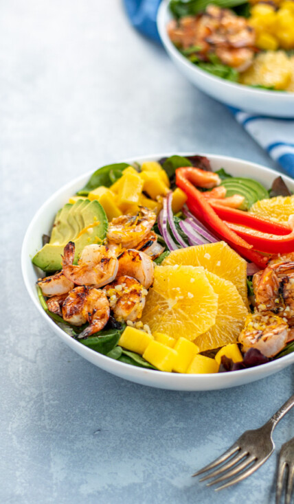 A large white bowl filled with mixed greens topped with grilled shrimp, avocado, oranges, mangos, red bell pepper, and red onions.