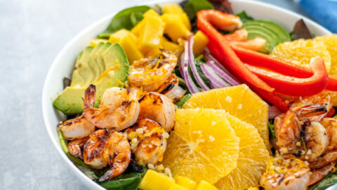 A large white bowl filled with mixed greens topped with grilled shrimp, avocado, oranges, mangos, red bell pepper, and red onions.