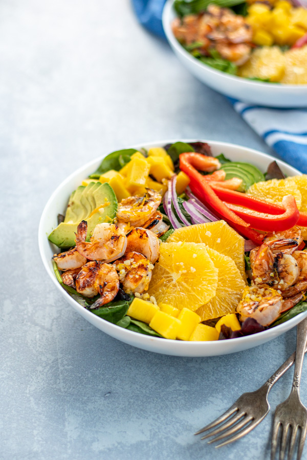 A large white bowl filled with mixed greens topped with grilled shrimp, avocado, oranges, mangos, red bell pepper, and red onions.