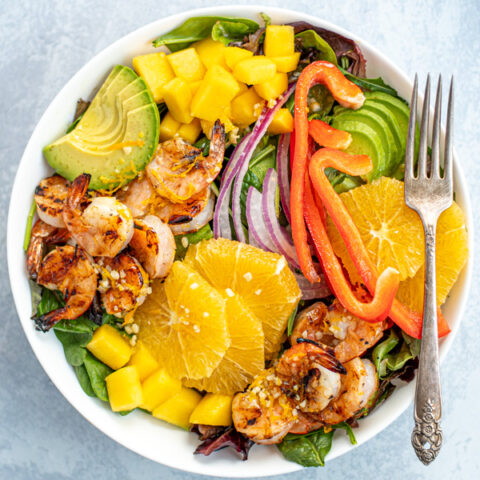 Overhead photo of a large white bowl filled with mixed greens with grilled shrimp, avocado, oranges, and mangos.