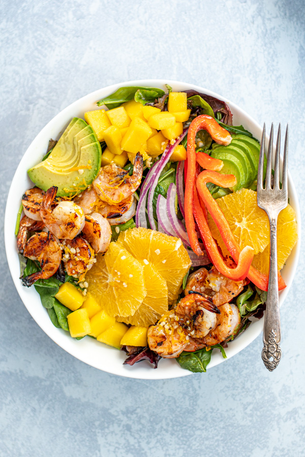 Overhead photo of a large white bowl filled with mixed greens with grilled shrimp, avocado, oranges, and mangos.