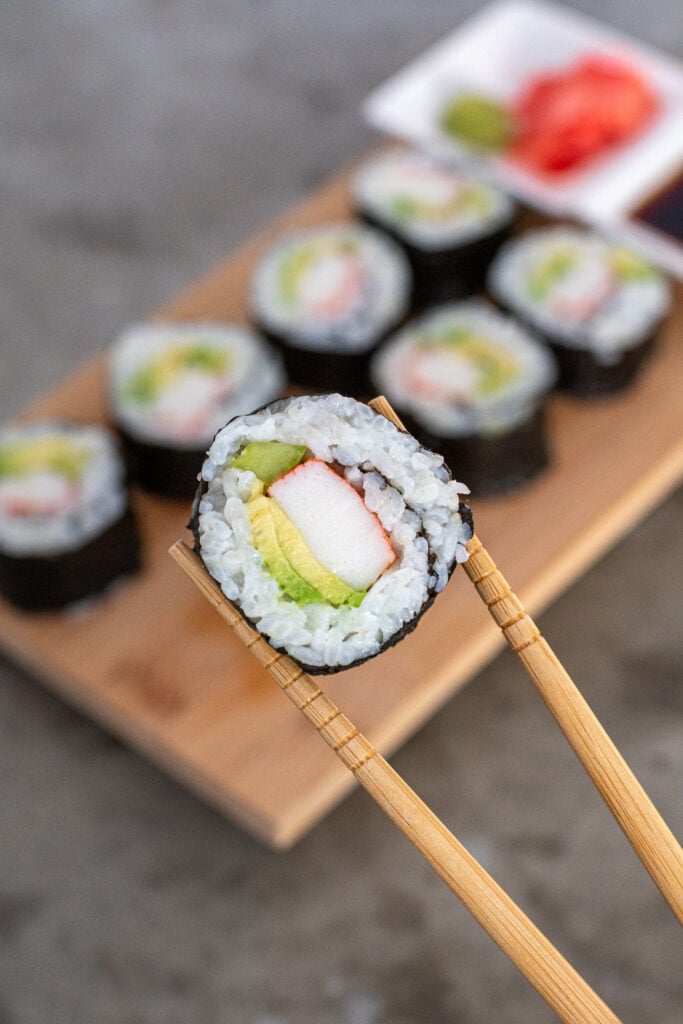 A pair of chopsticks holding up a California roll showing off the insides of crab, cucumber, and avocado.