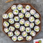 Large wooden platter filled with many California rolls next to some chopstick, soy sauce, and pink sushi ginger.