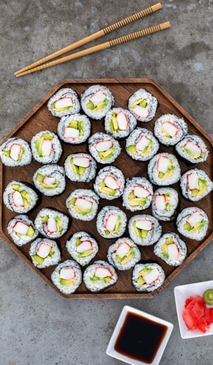 Large wooden platter filled with many California rolls next to some chopstick, soy sauce, and pink sushi ginger.