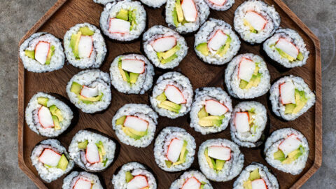 Large wooden platter filled with many California rolls next to some chopstick, soy sauce, and pink sushi ginger.