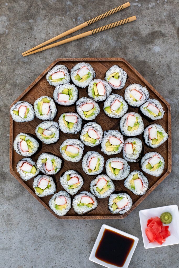 Large wooden platter filled with many California rolls next to some chopstick, soy sauce, and pink sushi ginger.