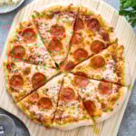 A cooked and cut pizza showing of how to make home pizza on a wooden pizza peel surrounded by ingredients.