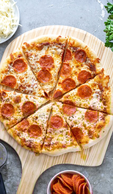 A cooked and cut pizza showing of how to make home pizza on a wooden pizza peel surrounded by ingredients.