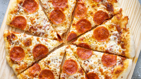 A cooked and cut pizza showing of how to make home pizza on a wooden pizza peel surrounded by ingredients.