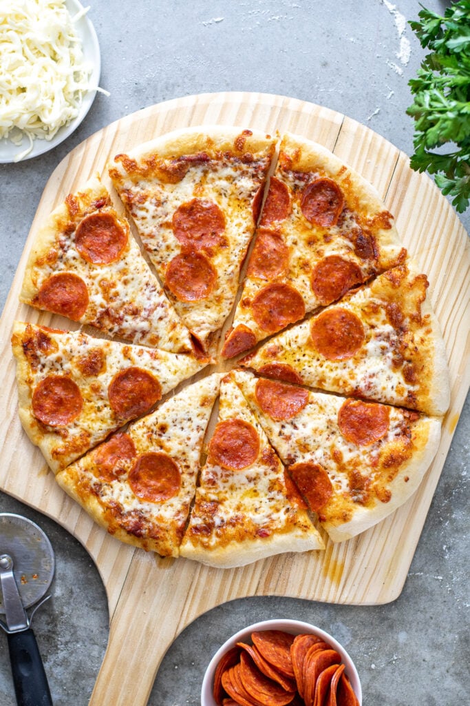 A cooked and cut pizza showing of how to make home pizza on a wooden pizza peel surrounded by ingredients.