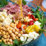 Pinterest Pin with text over lay 'Antipasto Salad with easy oregano dressing', image of a bowl of arugula topped with the ingredients of the salad with dressing being poured on.