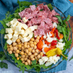 Pinterest Pin with text over lay 'Antipasto Salad with easy oregano dressing', image of a bowl of arugula topped with the ingredients of the salad.