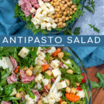 Pinterest Pin with text over lay 'Antipasto Salad', image of a bowl of arugula topped with the ingredients of the salad.