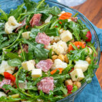 Pinterest Pin with text over lay 'Antipasto Salad with easy oregano dressing', image of a bowl of arugula topped with the ingredients of the salad.