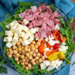 Pinterest Pin with text over lay 'Antipasto Salad', image of a bowl of arugula topped with the ingredients of the salad.